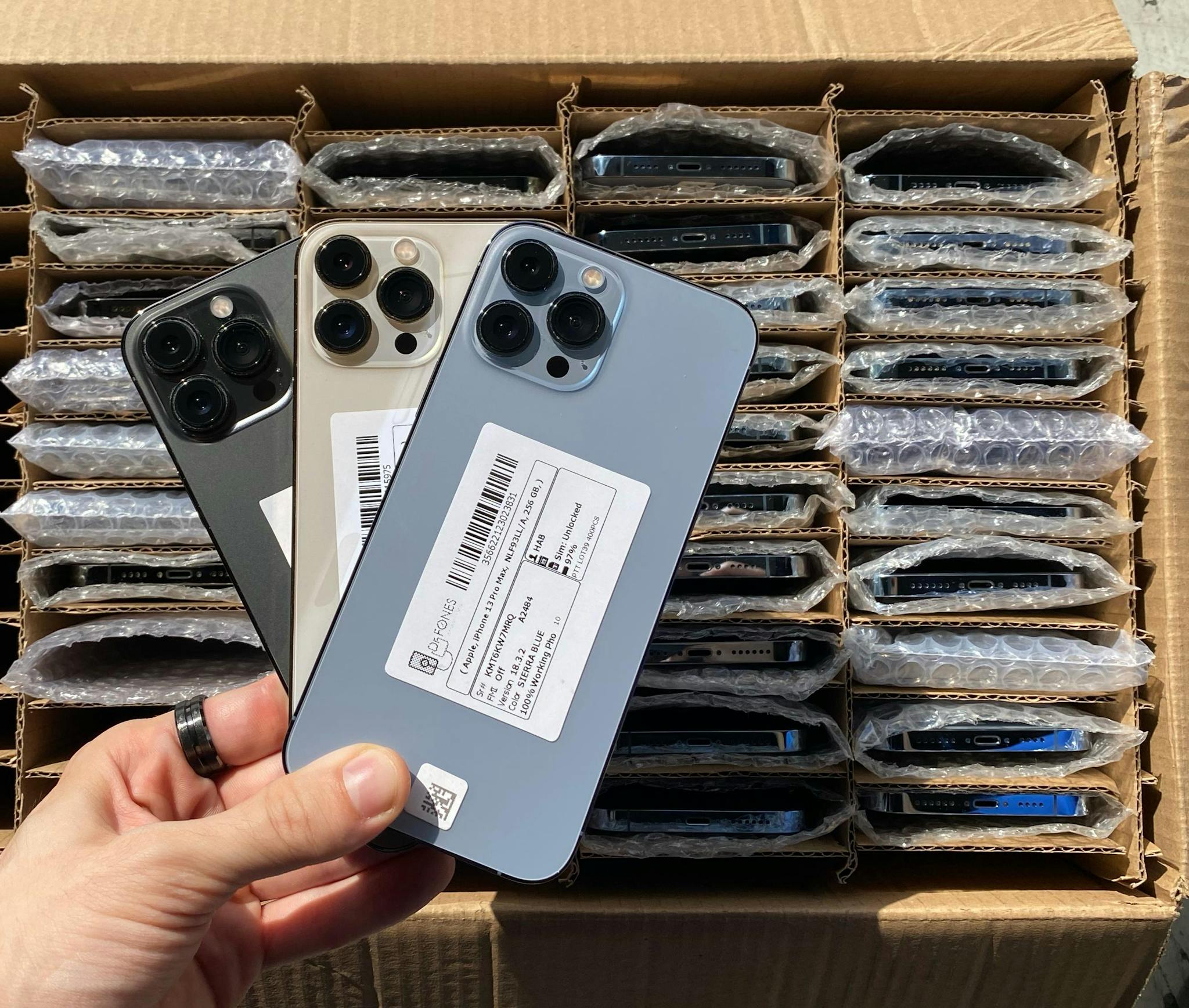 A hand holds three new smartphones over a box filled with packaged devices ready for shipment.
