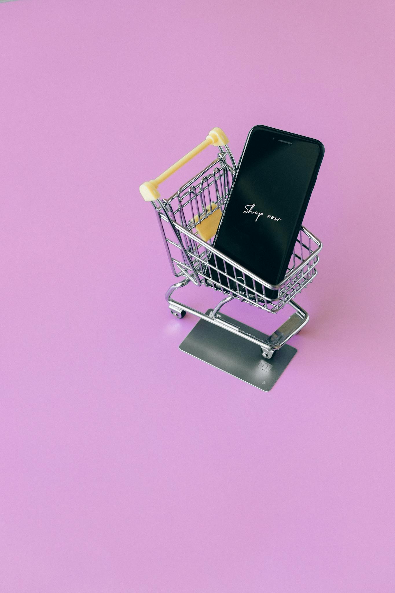 Mobile phone in mini cart on pink surface, symbolizing online shopping.
